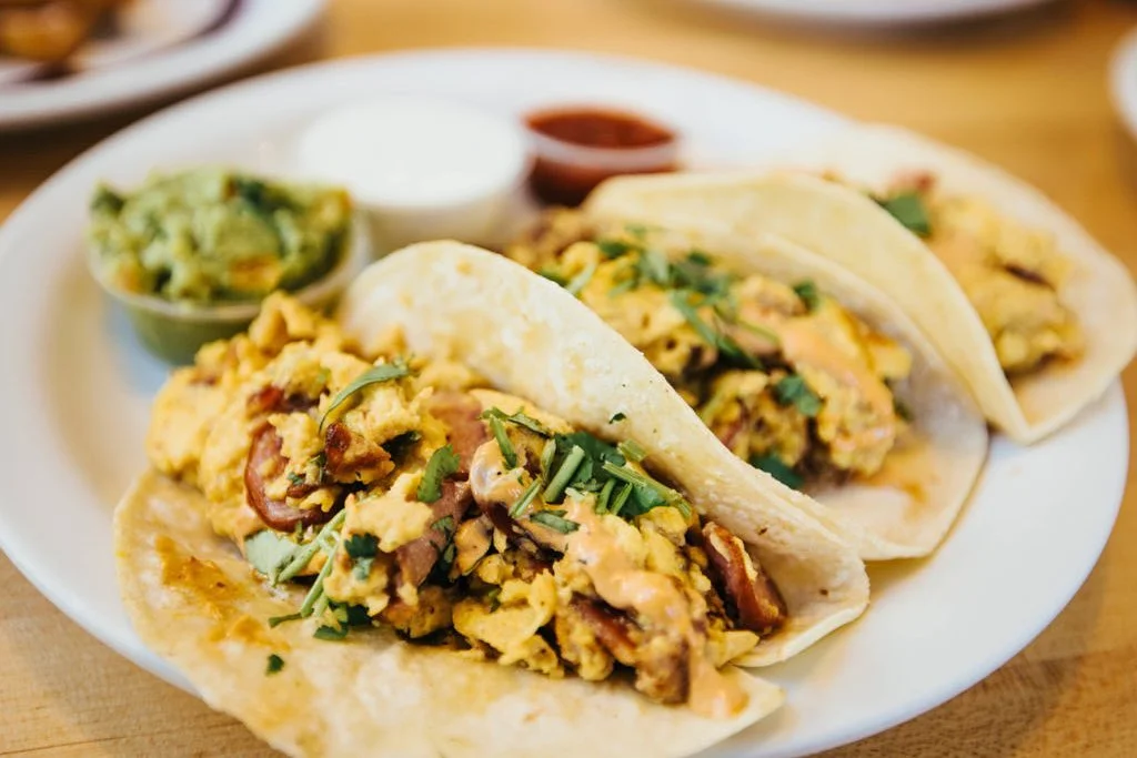 A plate of breakfast tacos filled with scrambled eggs, herbs, and meats, served with sides of guacamole, cream, and salsa.