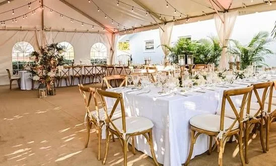 An elegant outdoor dining setup under a tent, featuring wooden chairs and tables adorned with tableware and floral arrangements.