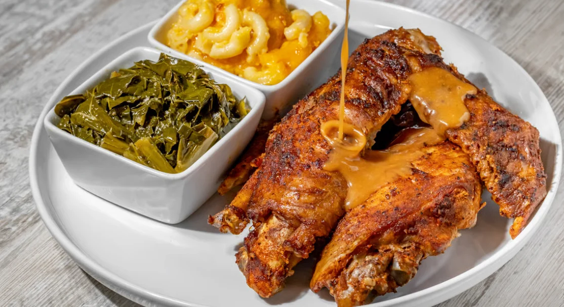 A plate featuring fried fish with gravy, collard greens, and macaroni and cheese in white bowls.