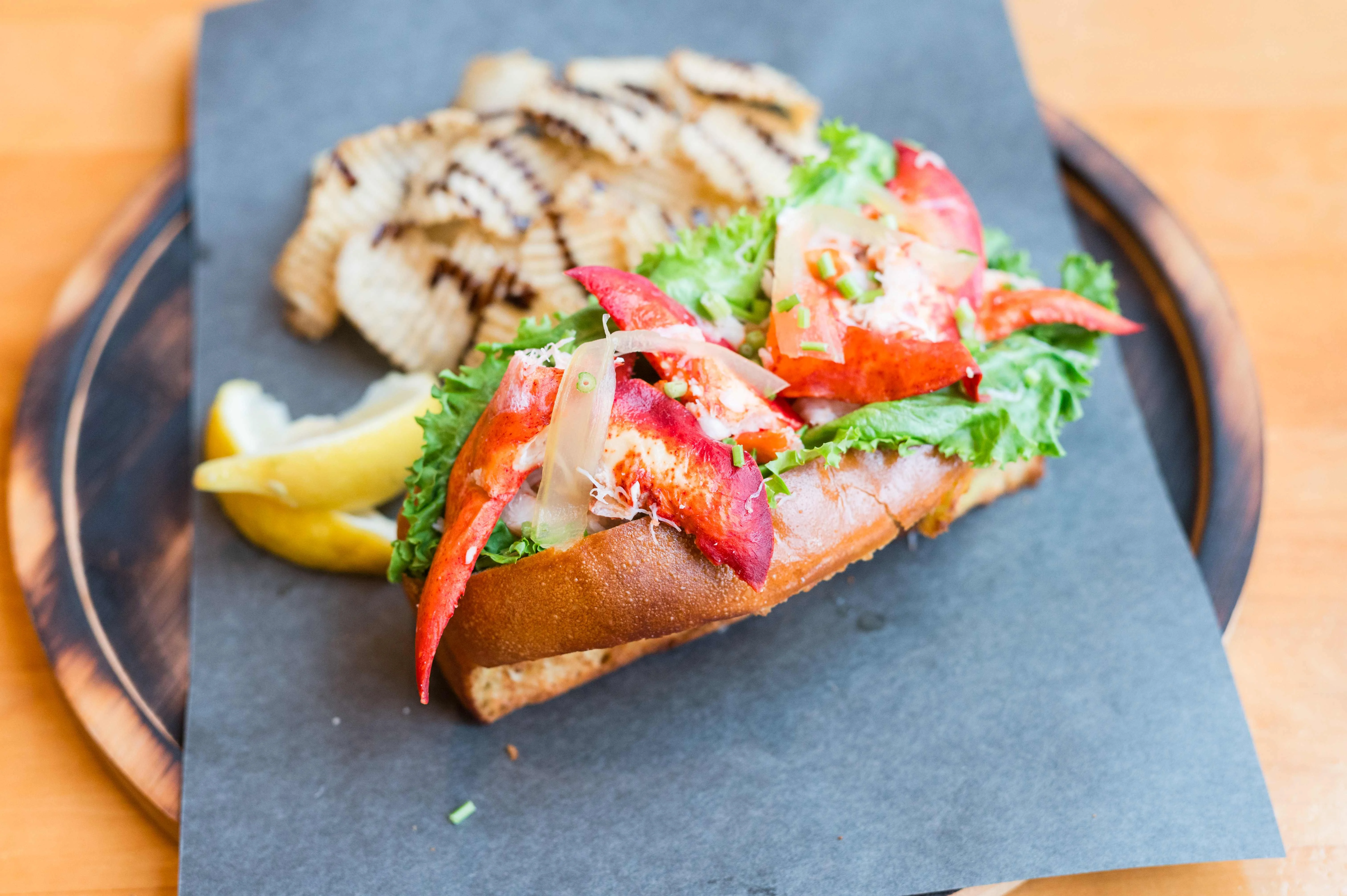 A lobster roll filled with chunks of lobster meat, served on a toasted bun with lettuce, accompanied by fried potato chips and a lemon wedge.