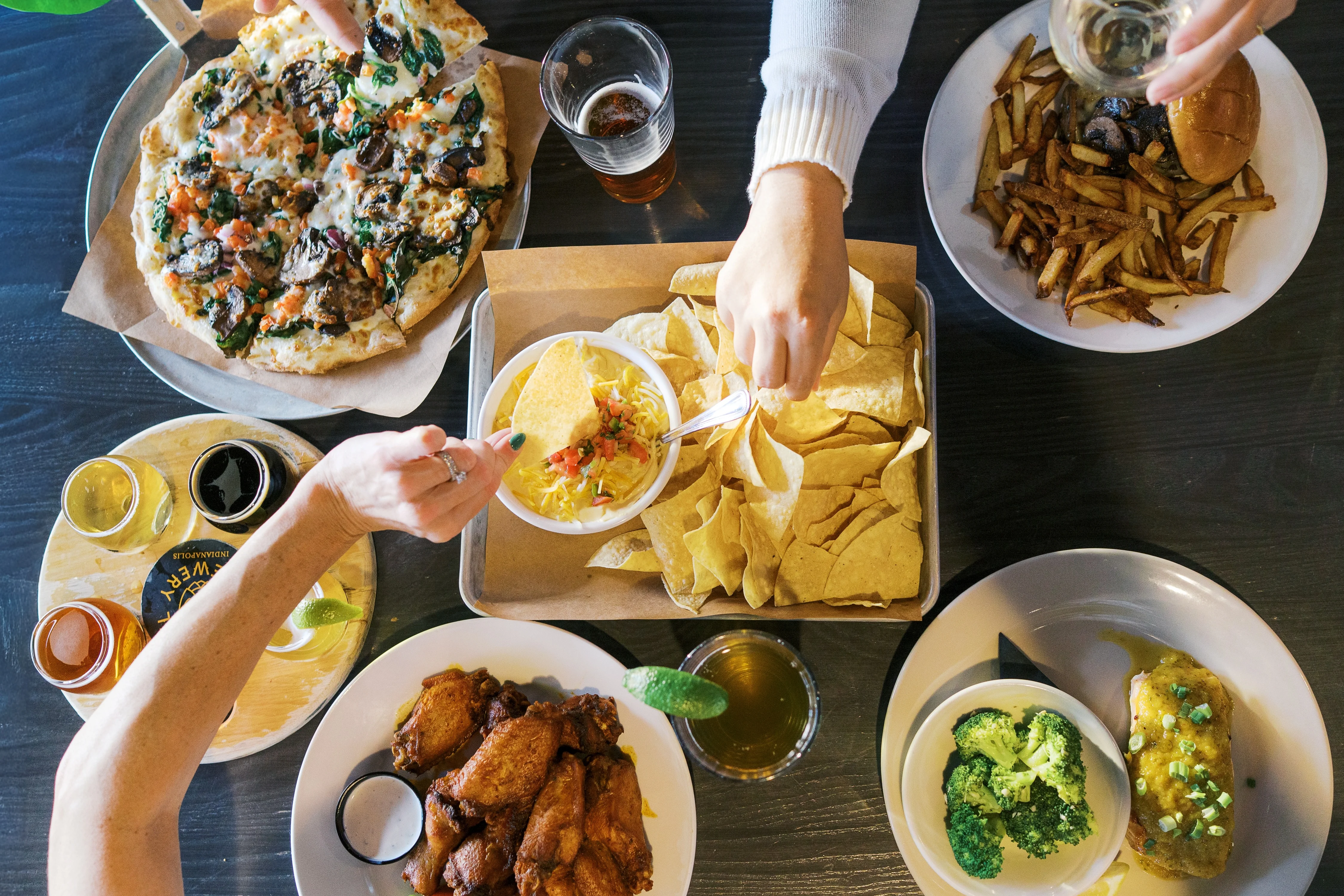 Overhead spread of restaurant dishes, nachos, wings, flatbread, sauces, and drinks
