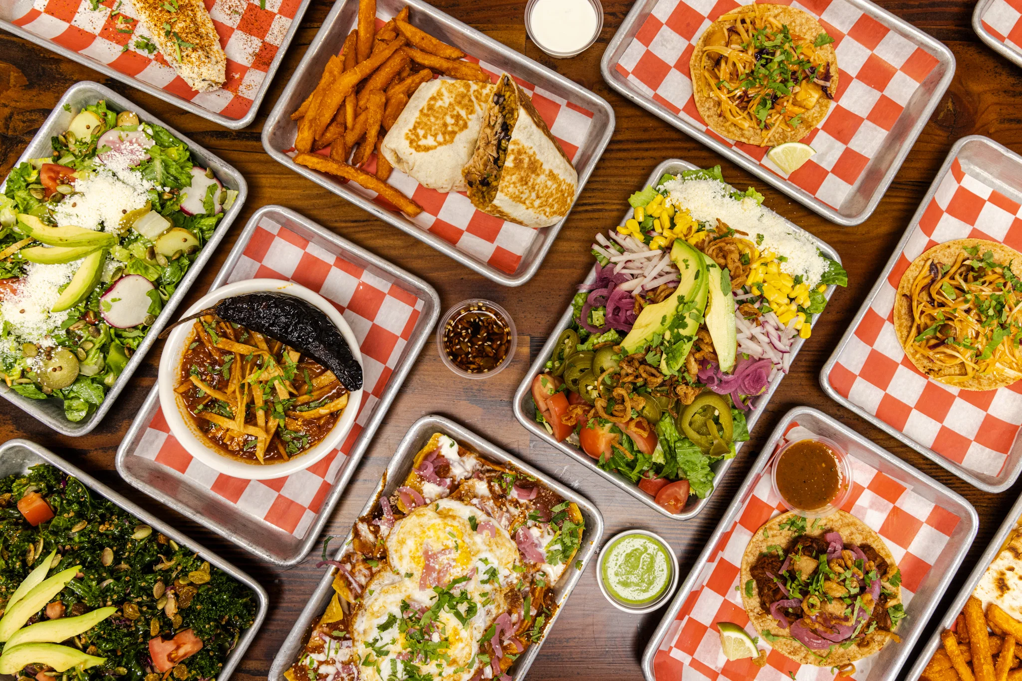An assortment of vibrant dishes including tacos, salads, and burritos served on metal trays. The meal features colorful vegetables, toppings, and dipping sauces.