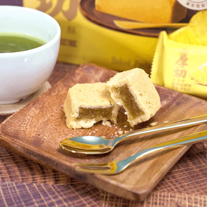 A wooden plate featuring cut pieces of a yellow cake, paired with a bowl of green tea. There are two golden spoons beside the cake.