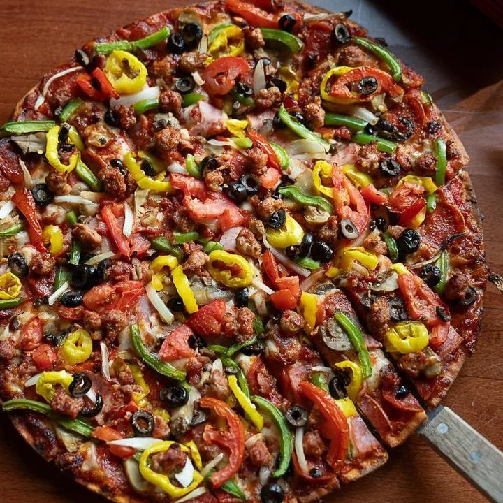 A spread of ingredients including tomatoes, green and yellow peppers, sausage, and slices of pepperoni, accompanied by a loaded pizza topped with various toppings.