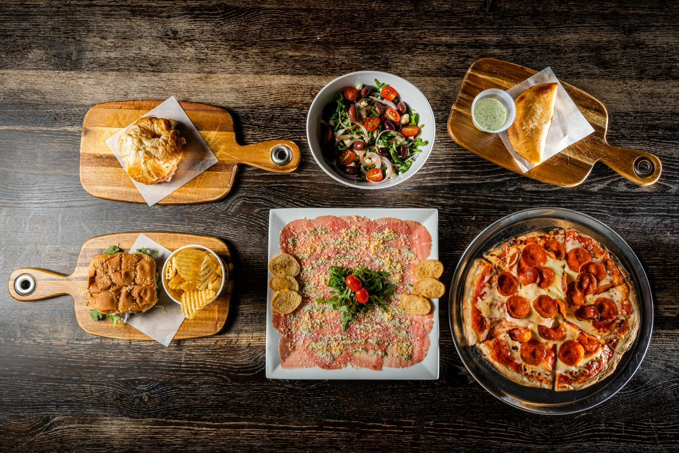 A variety of dishes arranged on wooden boards, including a salad, pizza, sashimi-style meat, and sandwiches, on a wooden table.