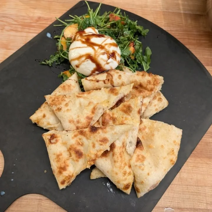 A serving of flatbread triangles arranged neatly on a dark plate, accompanied by a fresh salad and a scoop of creamy cheese drizzled with balsamic glaze.