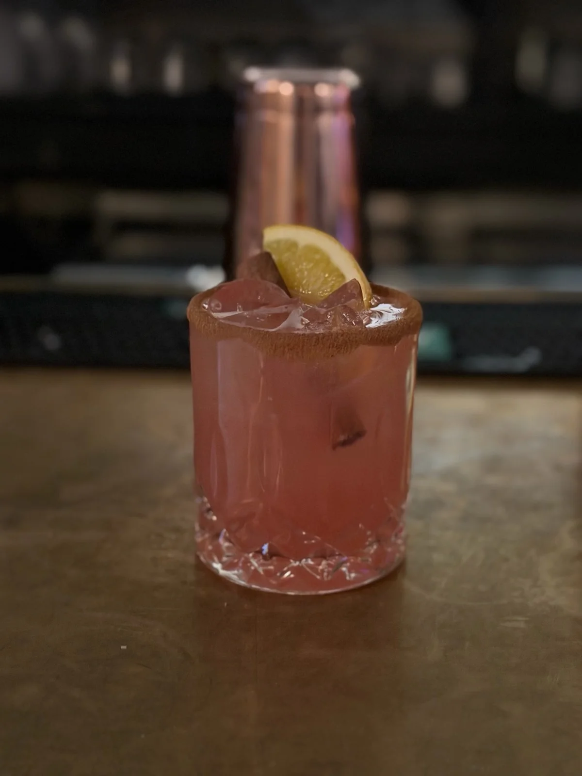 Pink cocktail with salted rim, ice, and lime wedge at a dimly lit bar