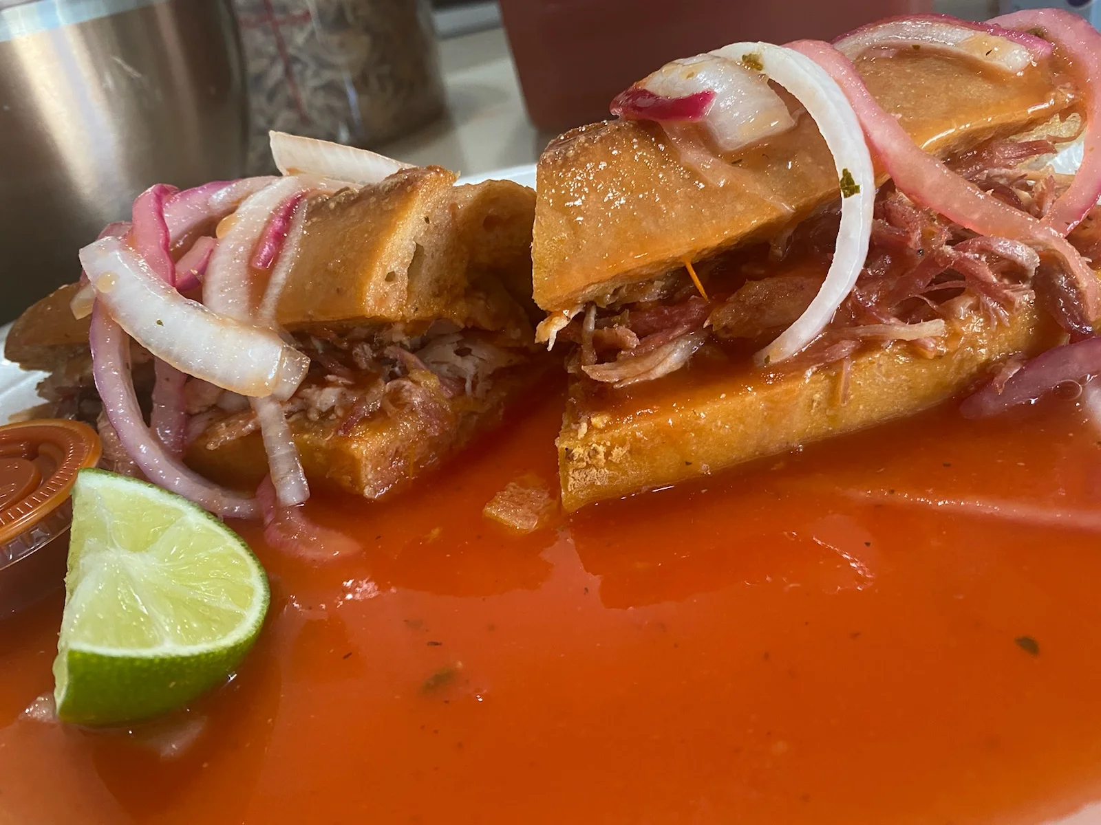 A delicious torta filled with shredded meat, topped with onions and served with a lime wedge and a side of sauce.
