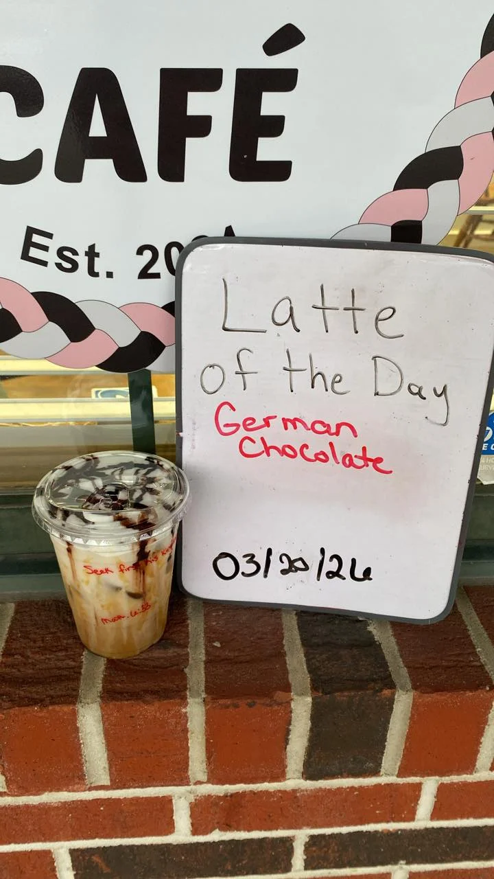 A sign advertising the latte of the day, which is German chocolate, next to a cup of iced coffee.