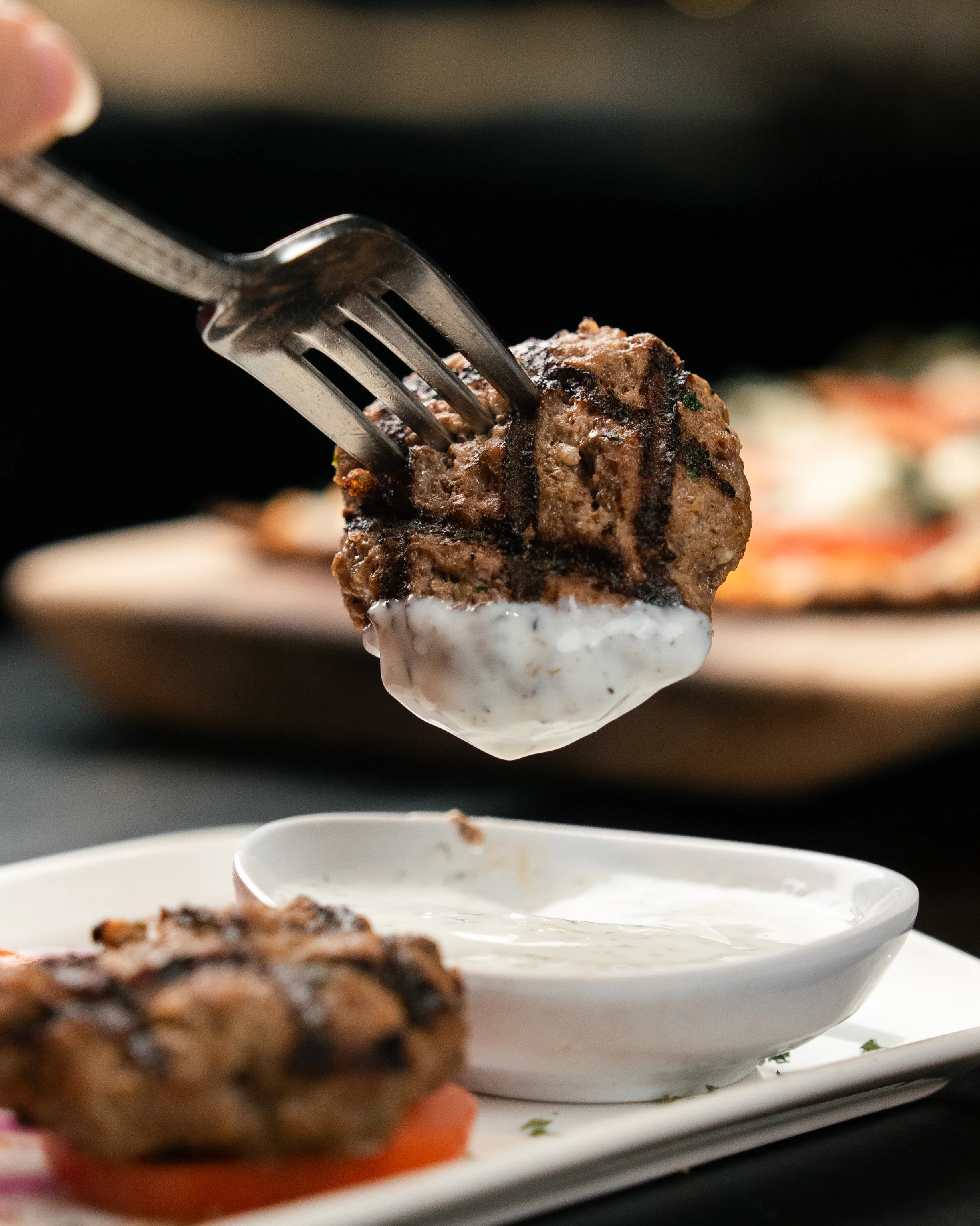 Grilled meatball being dipped in sauce with fresh vegetables on the side.