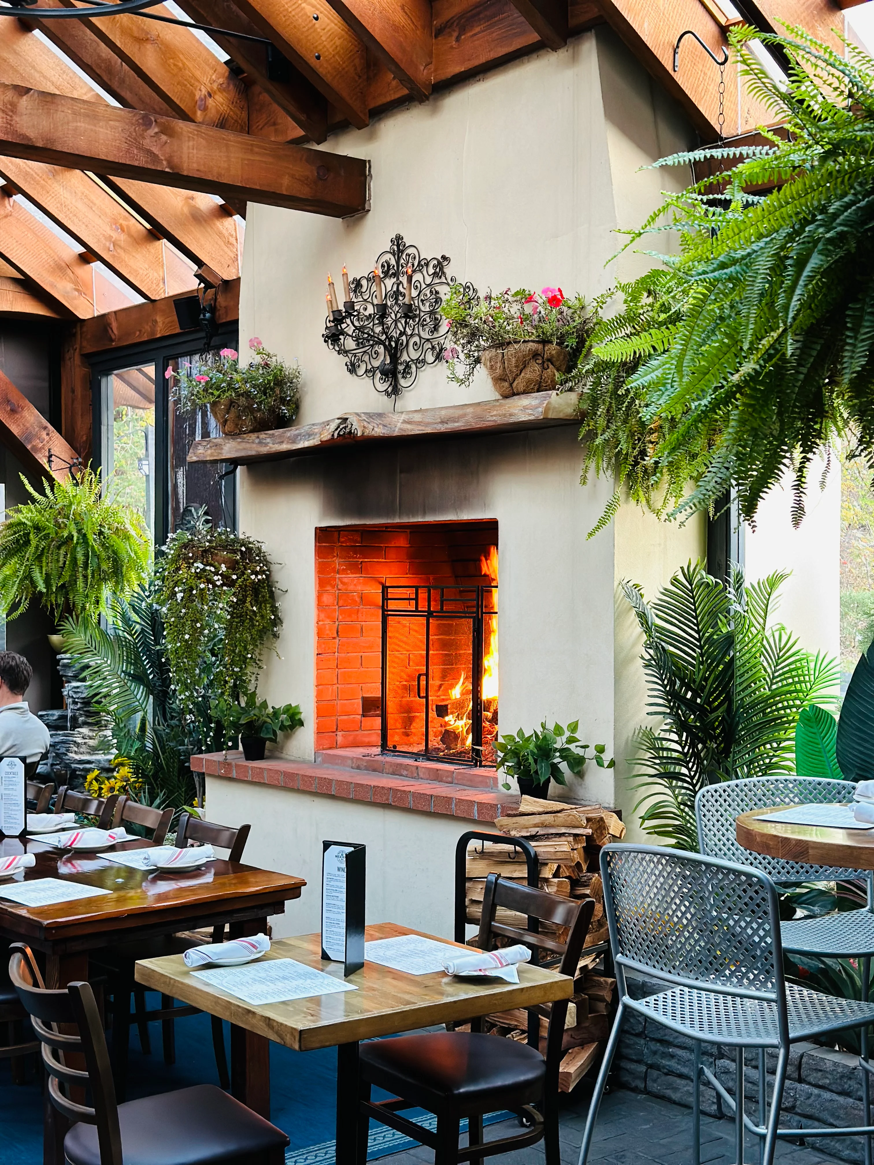A cozy indoor dining area featuring wooden tables, chairs, and a fireplace with a warm glow. The space is adorned with greenery and decorative elements, creating a welcoming atmosphere.