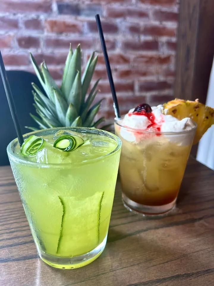 Two colorful cocktails placed on a wooden table with a pineapple in the background. One drink is green with cucumber slices, and the other has a fruit topping and whipped cream.