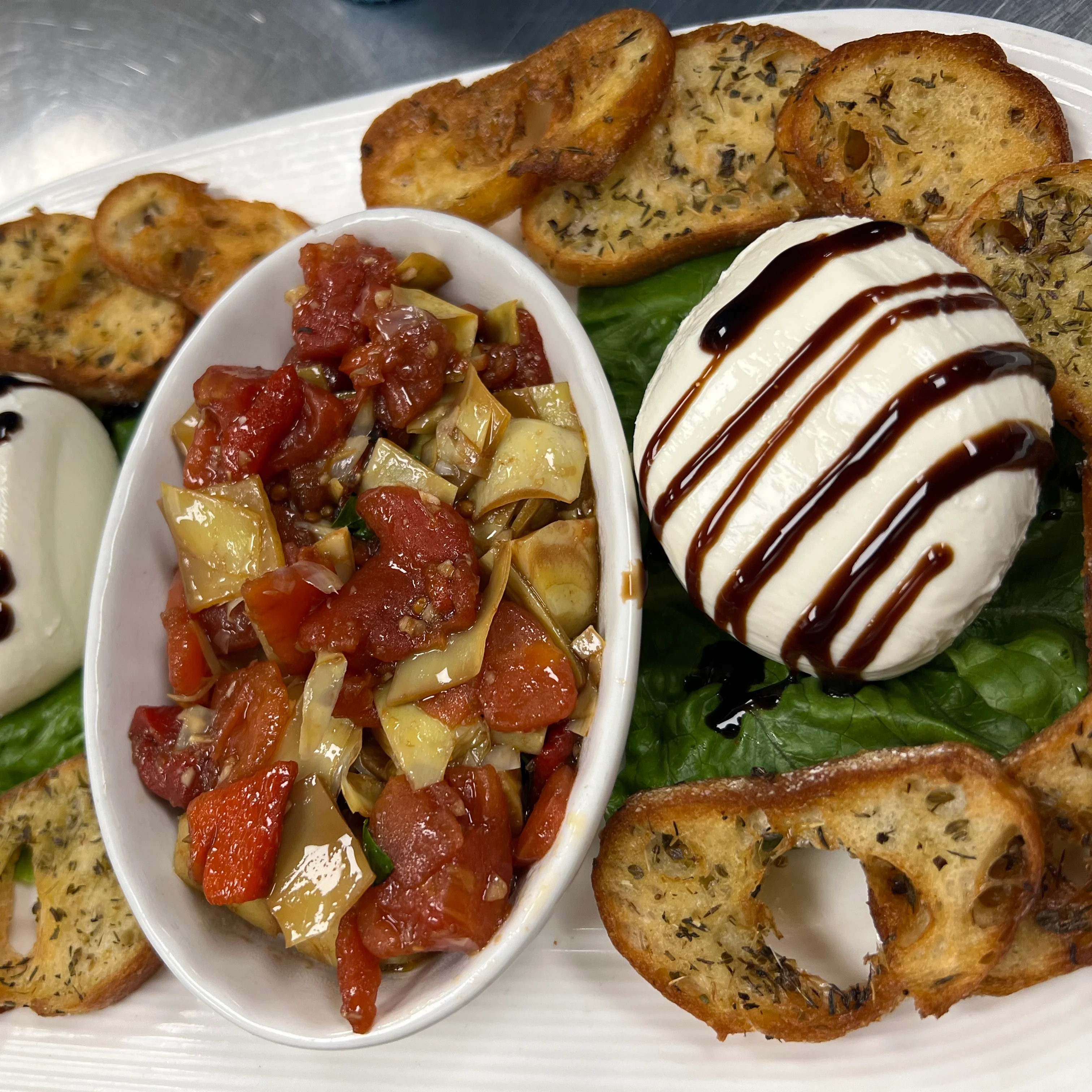 A beautifully plated appetizer featuring two balls of fresh mozzarella drizzled with balsamic glaze, surrounded by crispy herb crostini on a bed of green leaf lettuce. In the center is a small dish filled with a colorful tomato and artichoke bruschetta mix, creating a vibrant and appetizing presentation.