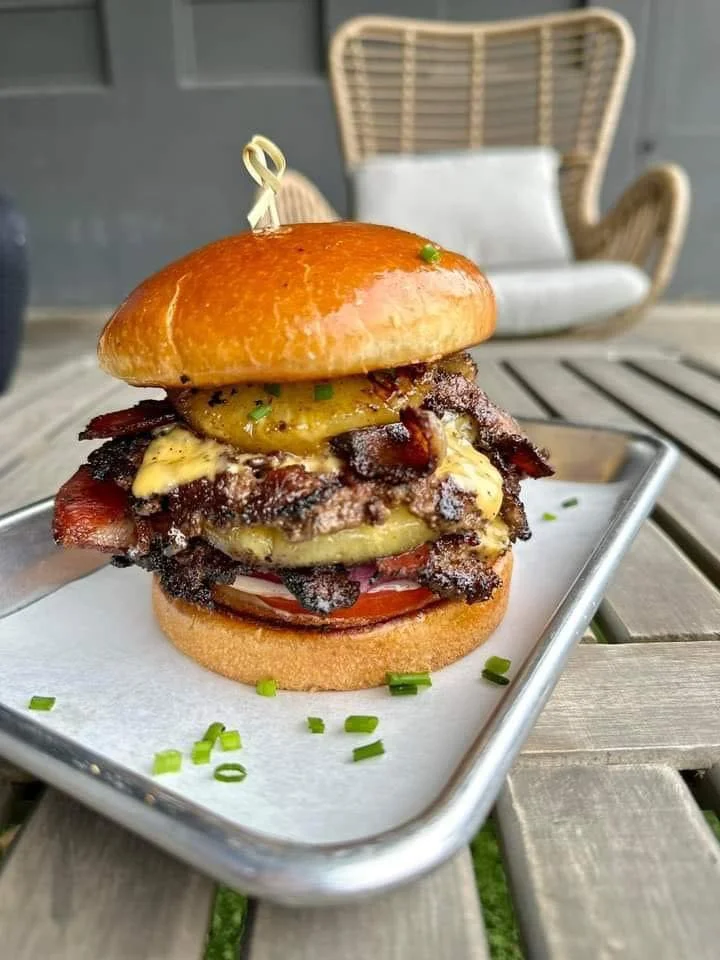 A gourmet burger stacked with beef, cheese, bacon, and grilled pineapple, served on a shiny brioche bun.