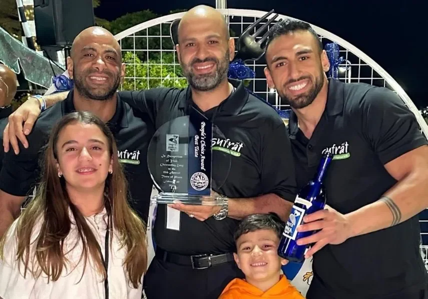 Three smiling men wearing black branded Sufrat shirts standing behind a little girl and a little boy. The man on the right is holding a blue wine bottle, and the man in the middle is holding a glass award plaque. The three men are the founders of Sufrat.