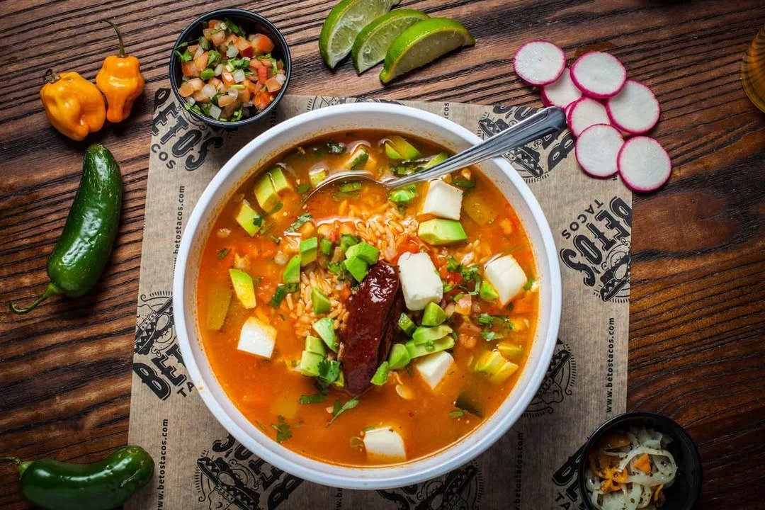 A bowl of colorful soup with vegetables, rice, and toppings, served with sides.