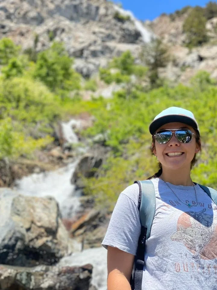 A hiker smiling in front of a lush green landscape with a waterfall in the background.
