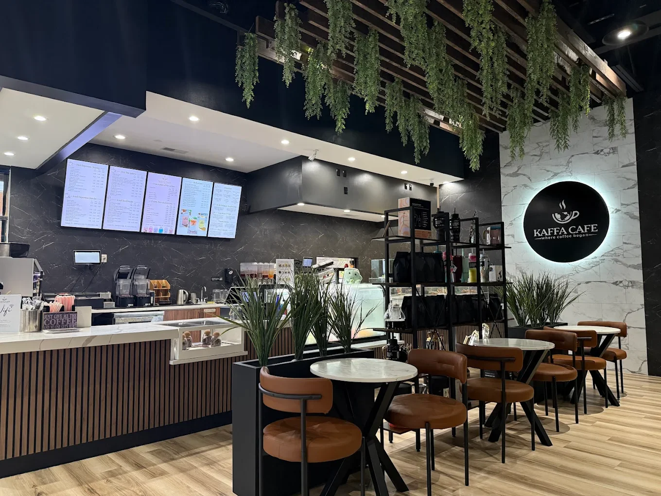 A modern café interior featuring a counter with menu screens, seating areas with round tables and brown chairs, and decorative plants hanging from the ceiling.