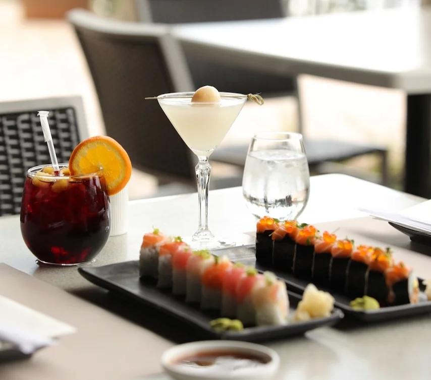 An outdoor setting featuring two platters of specialty sushi rolls, a lychee martini, and a dark berry cocktail garnished with an orange slice.