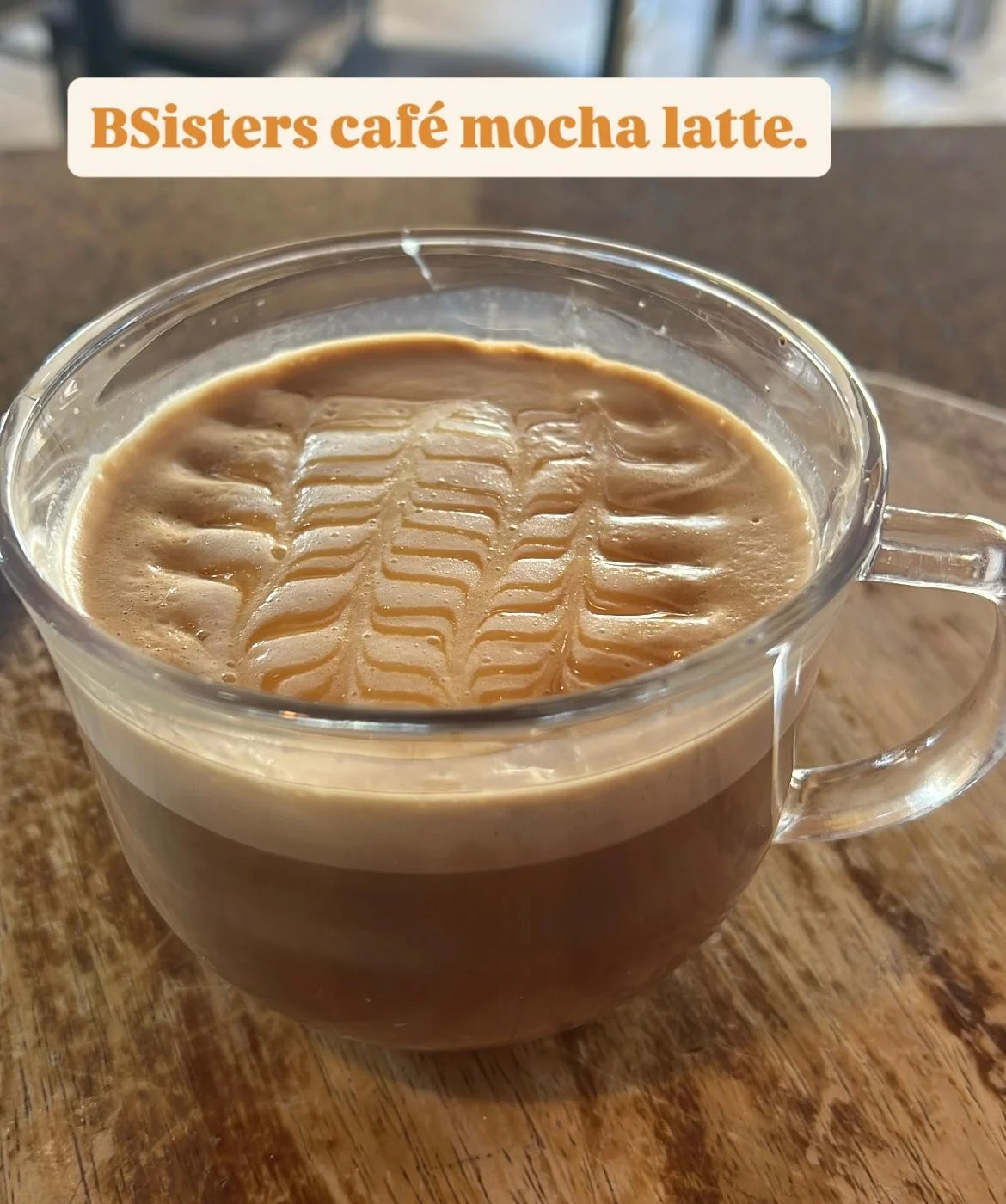 A clear cup of mocha latte featuring a beautifully crafted foam design on top, sitting on a wooden table.
