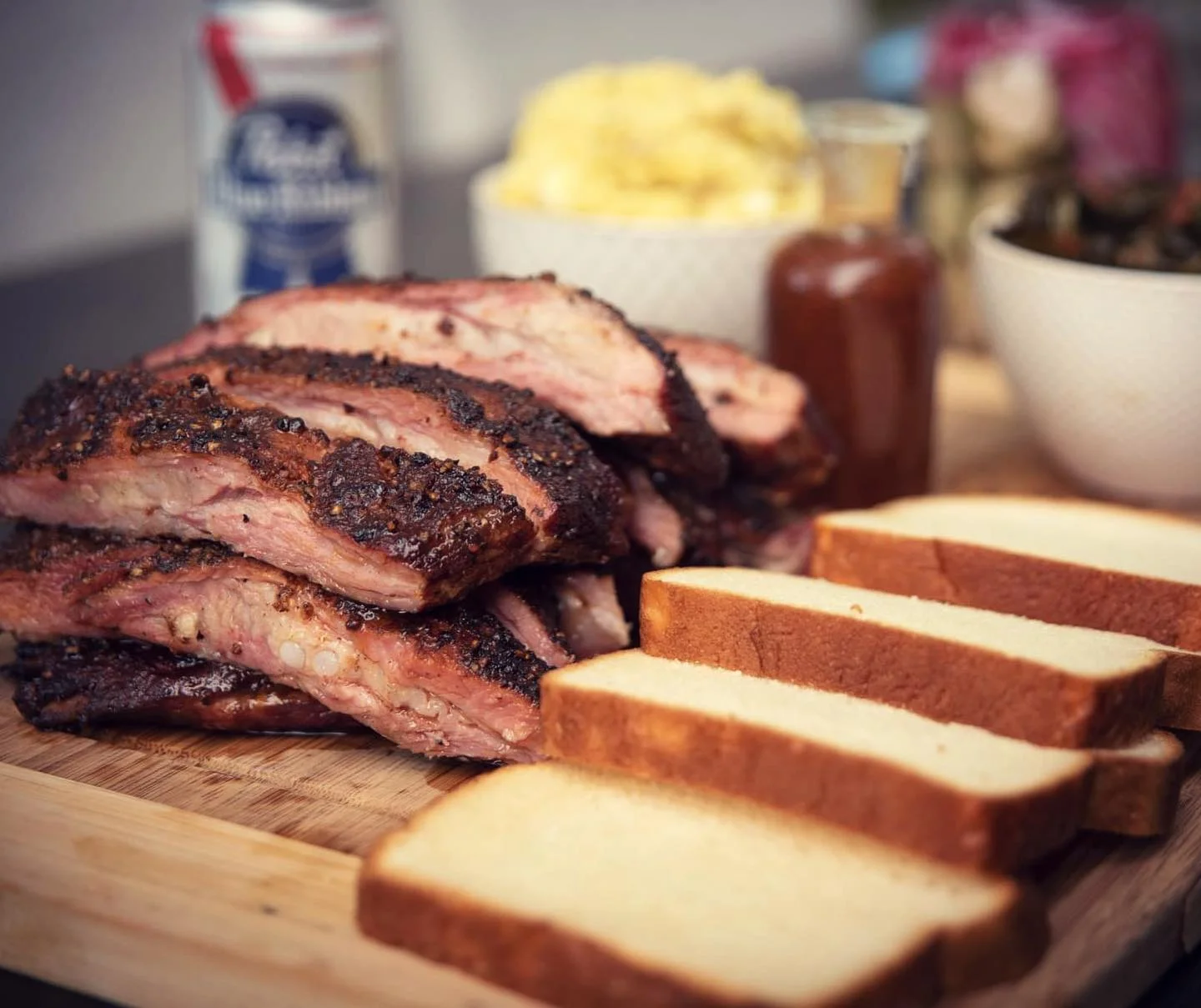 A platter of barbecued ribs alongside slices of bread, mashed potatoes, and a small bowl of greens.