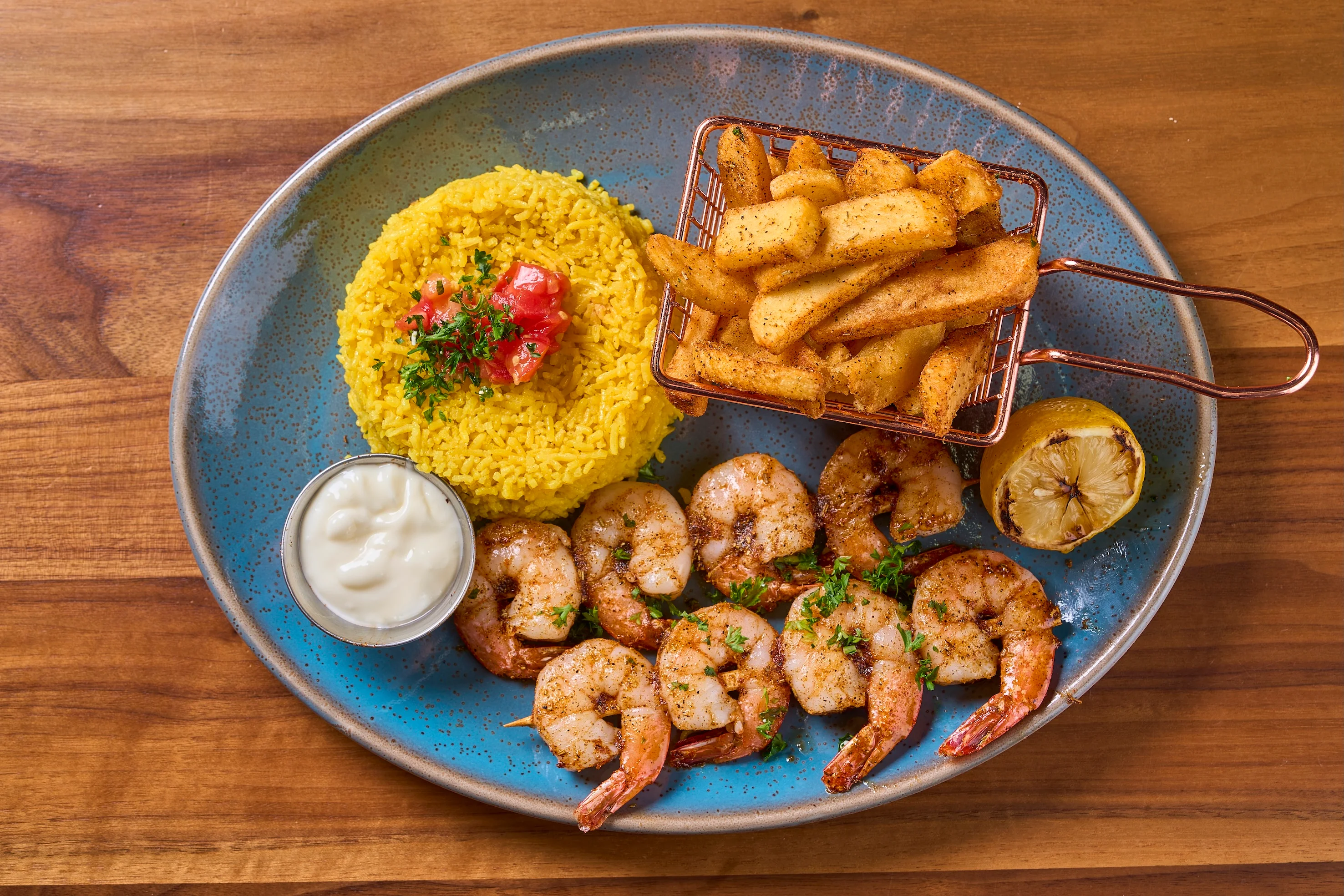 A blue plate with grilled shrimp garnished with herbs, a mound of yellow rice with tomato, a basket of seasoned fries, aioli, and a grilled lemon half.