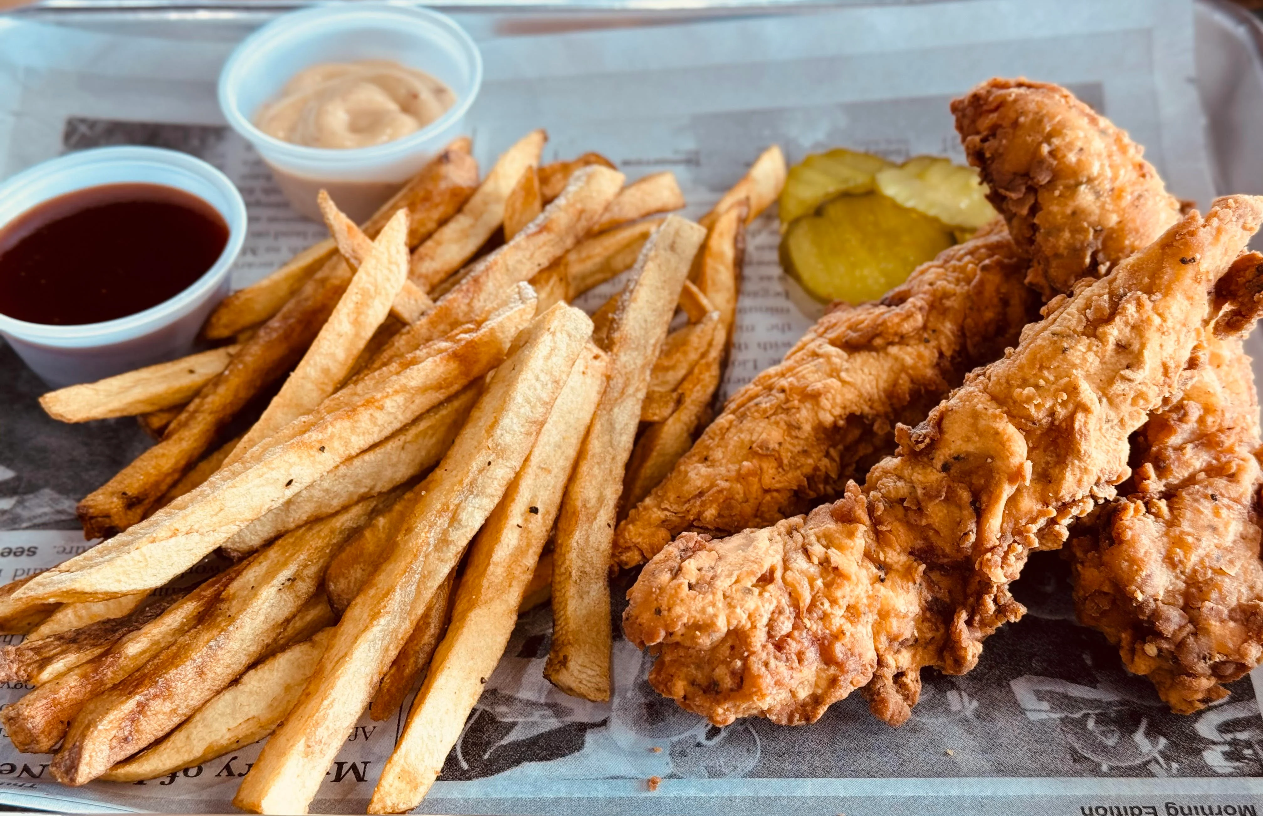 A platter featuring fried chicken tenders with French fries and dipping sauces, accompanied by pickles.