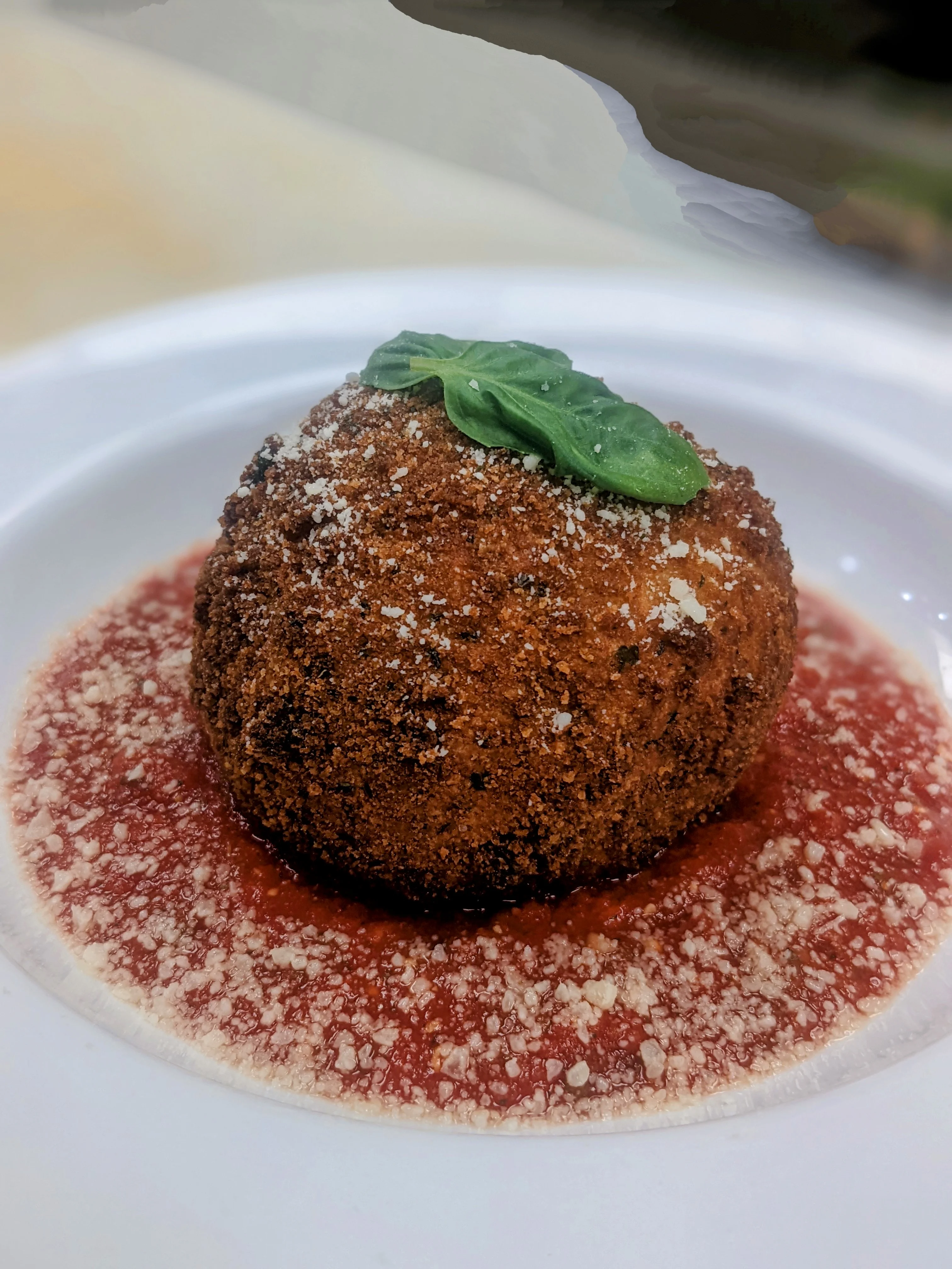 A breaded and fried rice ball served on a pool of marinara sauce, garnished with grated cheese and a basil leaf.