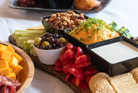 A colorful platter of snacks featuring a variety of vegetables, dips, nuts, and cheese. There are bowls with cheese spread, olives, and diced veggies, complemented by celery and crackers.