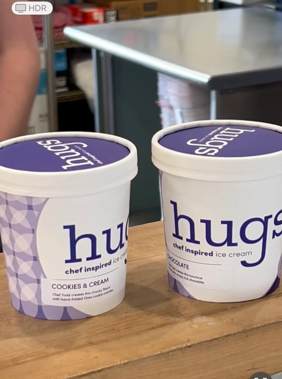 Two pints of Hugs chef inspired ice cream on a wooden counter—one labeled Cookies & Cream with a light purple patterned design, and the other labeled Chocolate with a solid white label and purple lid—set in a commercial kitchen environment.