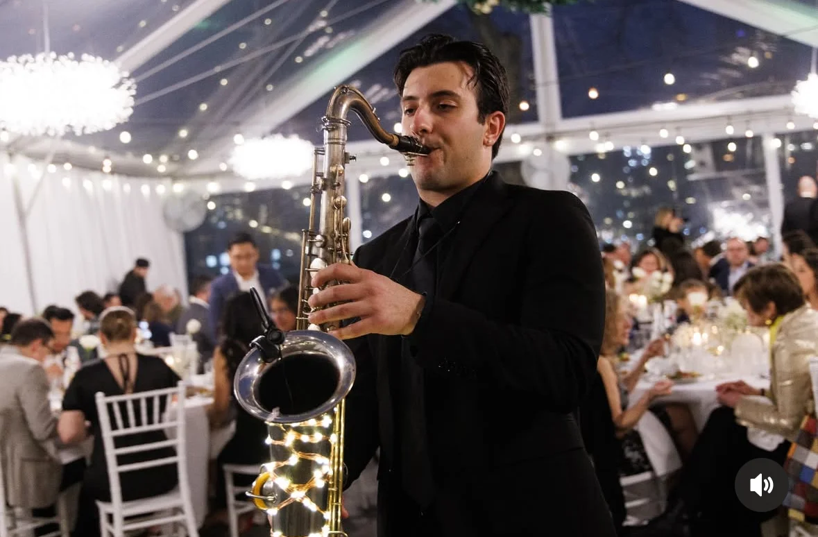 A musician playing a saxophone at a festive indoor event with tables set for guests and soft lighting.
