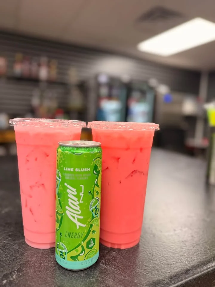 Two pink drinks with ice beside a green energy drink can, arranged on a dark counter with a blurred background.