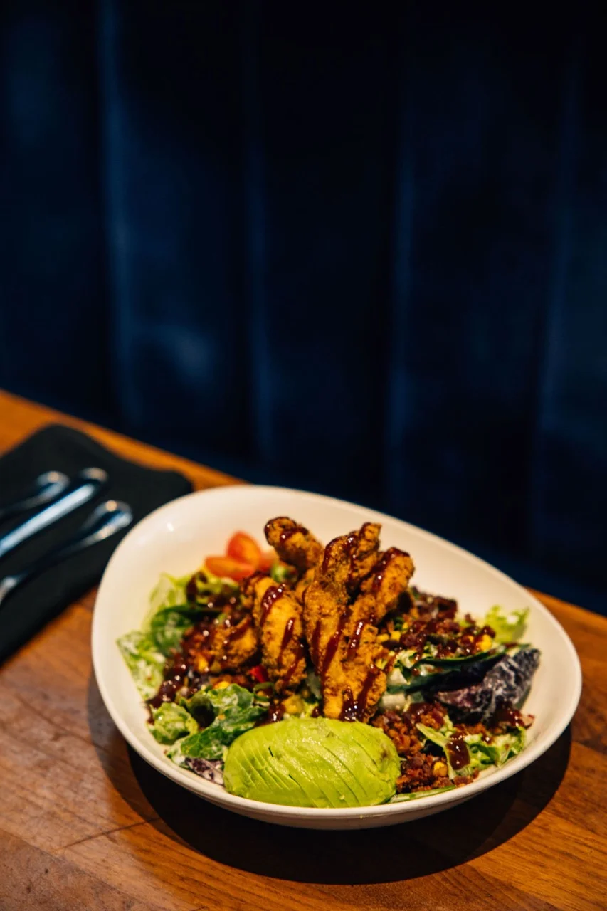 A colorful salad featuring crispy chicken, greens, and avocado, accompanied by a few cherry tomatoes, beautifully arranged on a wooden table.