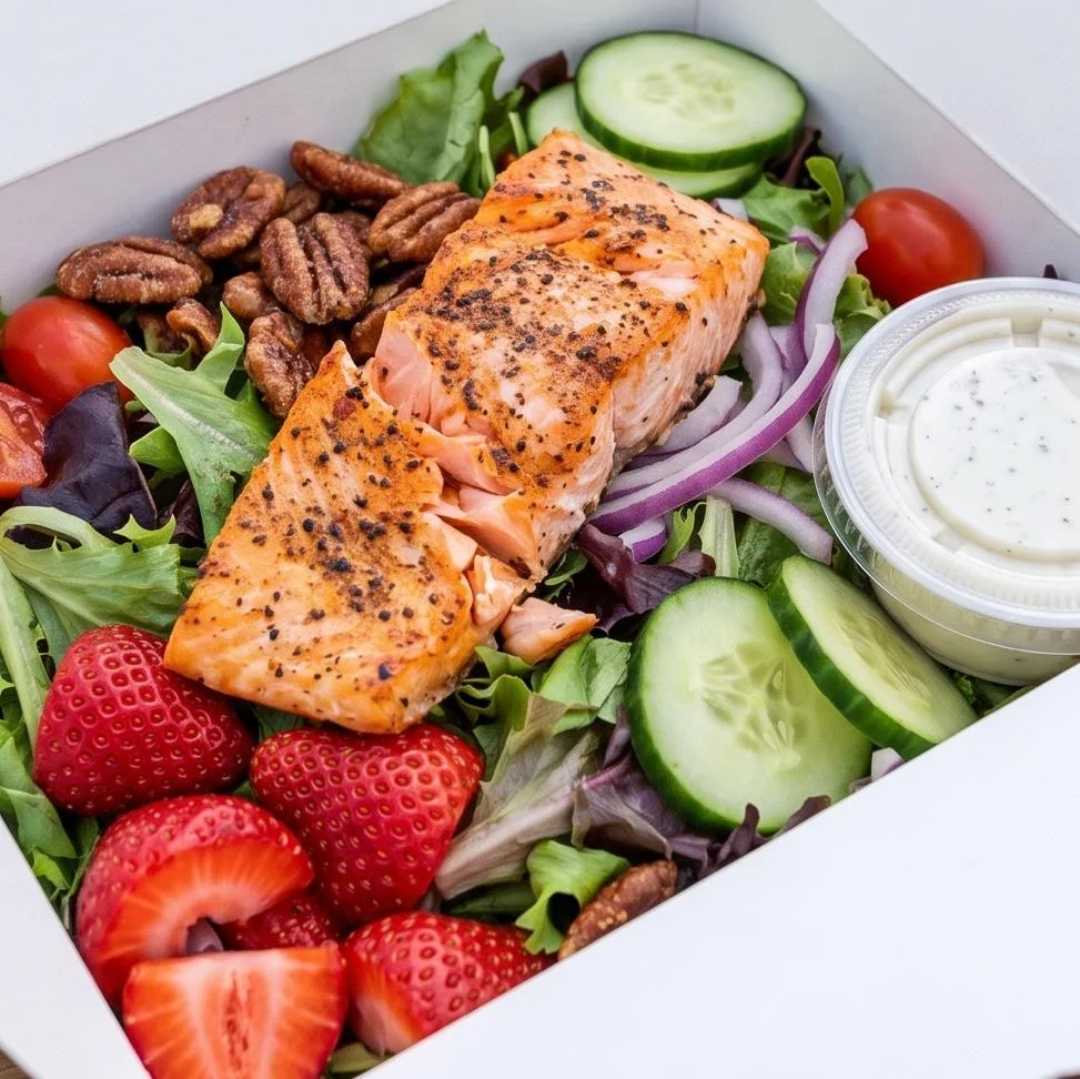 A healthy salad featuring grilled salmon, fresh greens, strawberries, cucumbers, and a side of dressing.