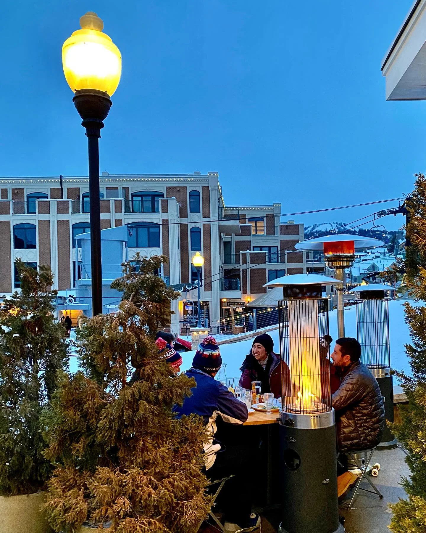 A group of people gathered around a table outside, with a gas patio heater providing warmth.