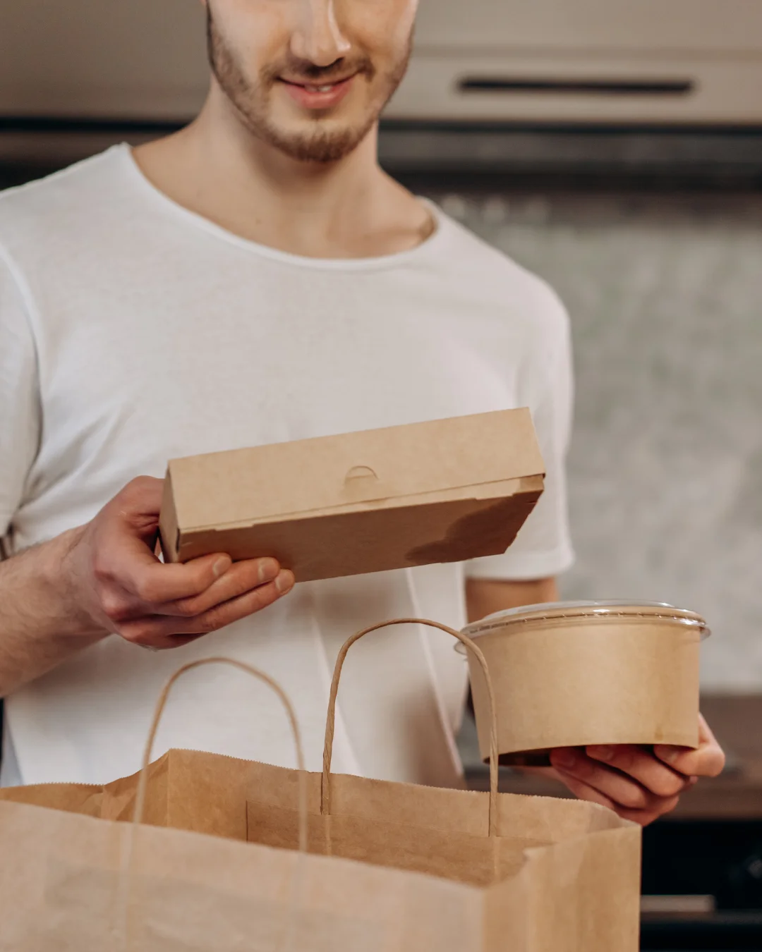 Smiling man in a white t-shirt holding takeout containers—one rectangular and one round with a clear lid—above a paper grocery bag, in a home kitchen setting.