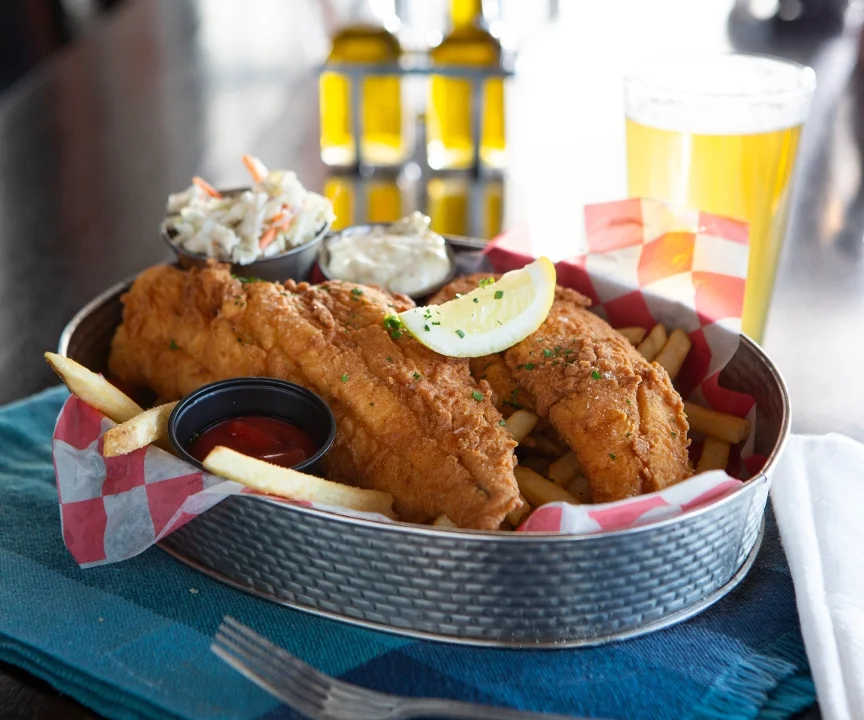 A platter featuring two pieces of fried fish, French fries, coleslaw, and sauces, garnished with a slice of lemon.
