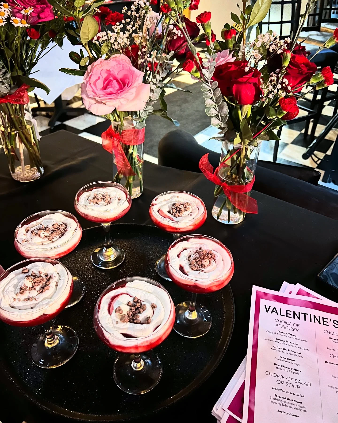 A Valentine's Day dessert spread featuring six elegant glasses of whipped dessert topped with chocolate bits, arranged on a black serving tray. The table is adorned with vases of red and pink roses, carnations, and baby's breath, wrapped in red ribbon. A stack of Valentine's Day menus is placed on the side, creating a romantic and festive ambiance.