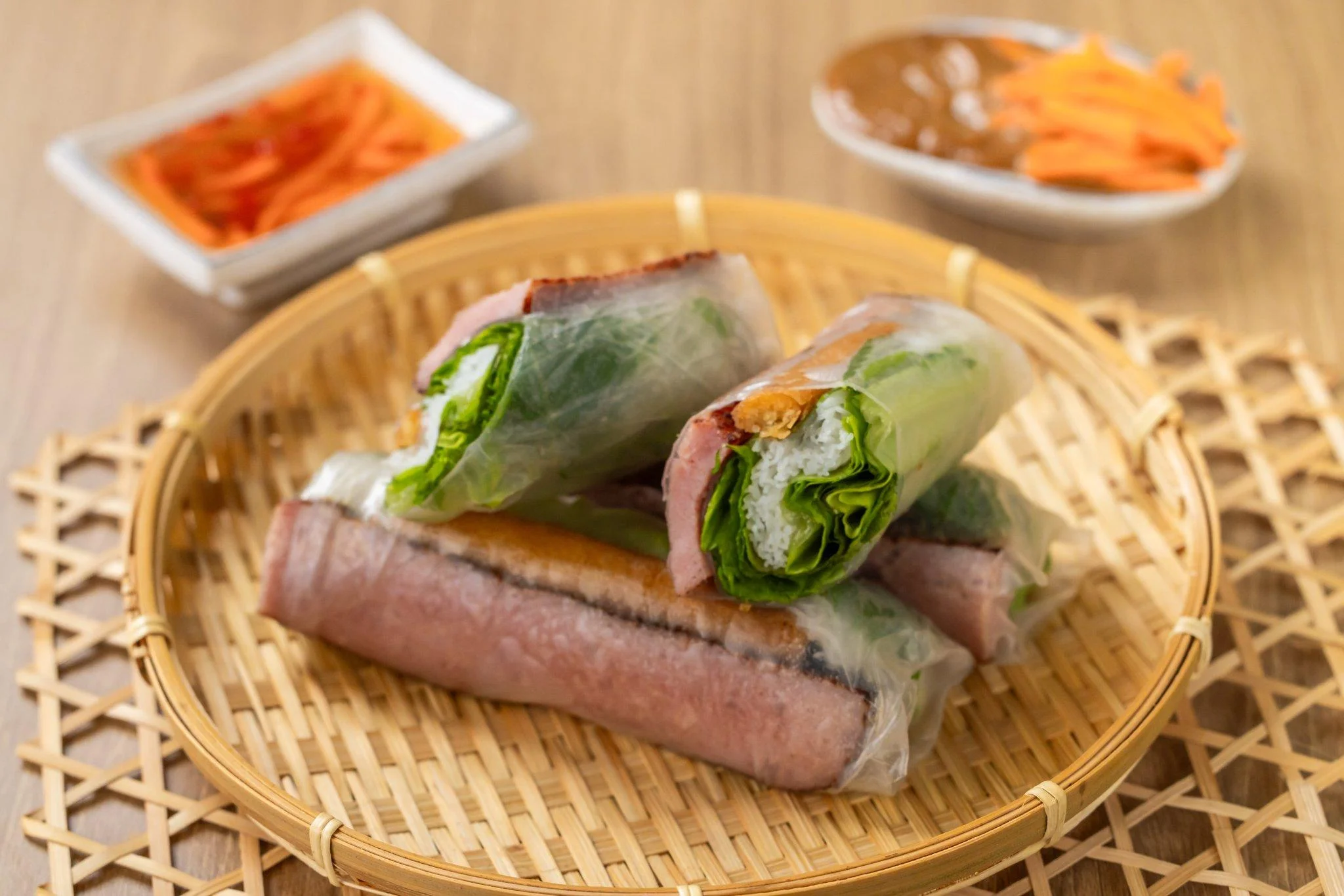 A bamboo basket with spring rolls containing greens and another ingredient, accompanied by a side of pickled carrots and dipping sauce.