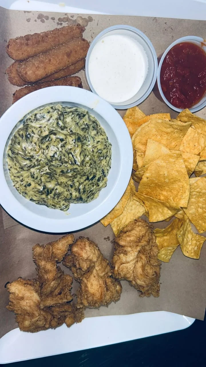 A platter featuring fried chicken, spinach dip, crispy tortilla chips, and some cheese sticks, served with dipping sauces.