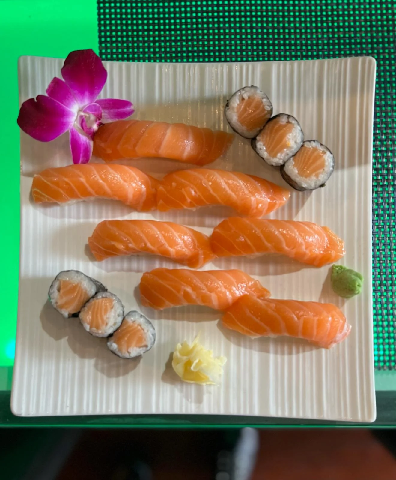 A beautifully arranged plate of sushi featuring several pieces of salmon nigiri, accompanied by salmon rolls, wasabi, pickled ginger, and an orchid flower.