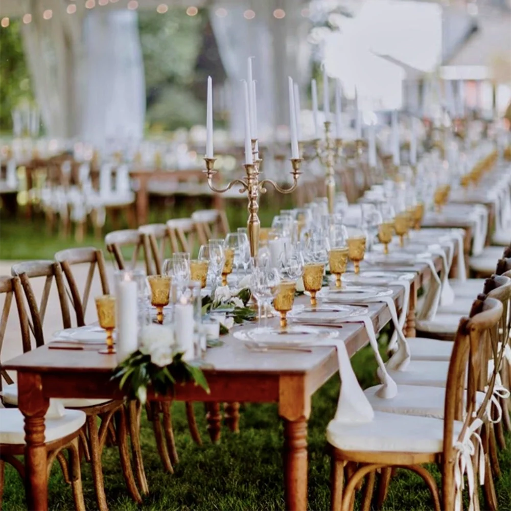 An elegantly decorated outdoor dining setting with long wooden tables, adorned with candle holders and glassware, set on a lawn.