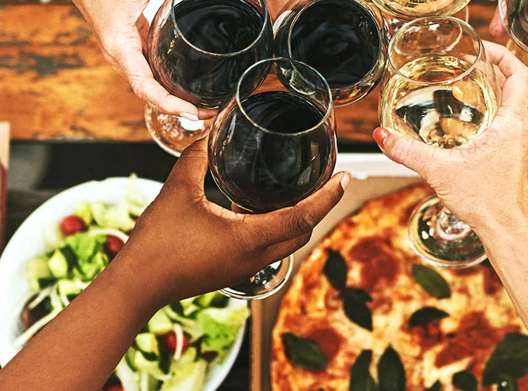 People clinking wine glasses over a table with pizza and salad on it.