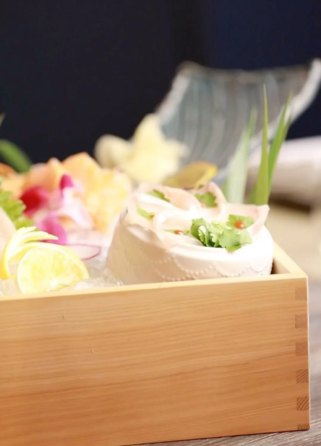 An elegant sashimi presentation arranged inside a light wooden box filled with ice, highlighting thin slices of white fish shaped like a flower in a small white ceramic dish, garnished with fresh cilantro and dots of red sauce.