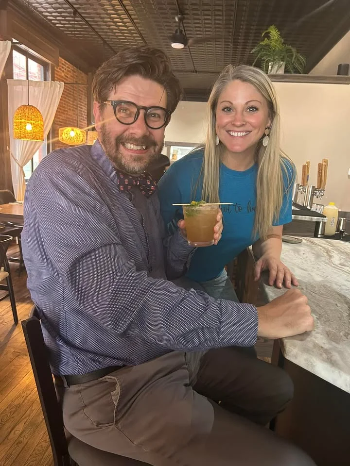 Two people posing together in a casual dining setting, enjoying drinks and smiling.