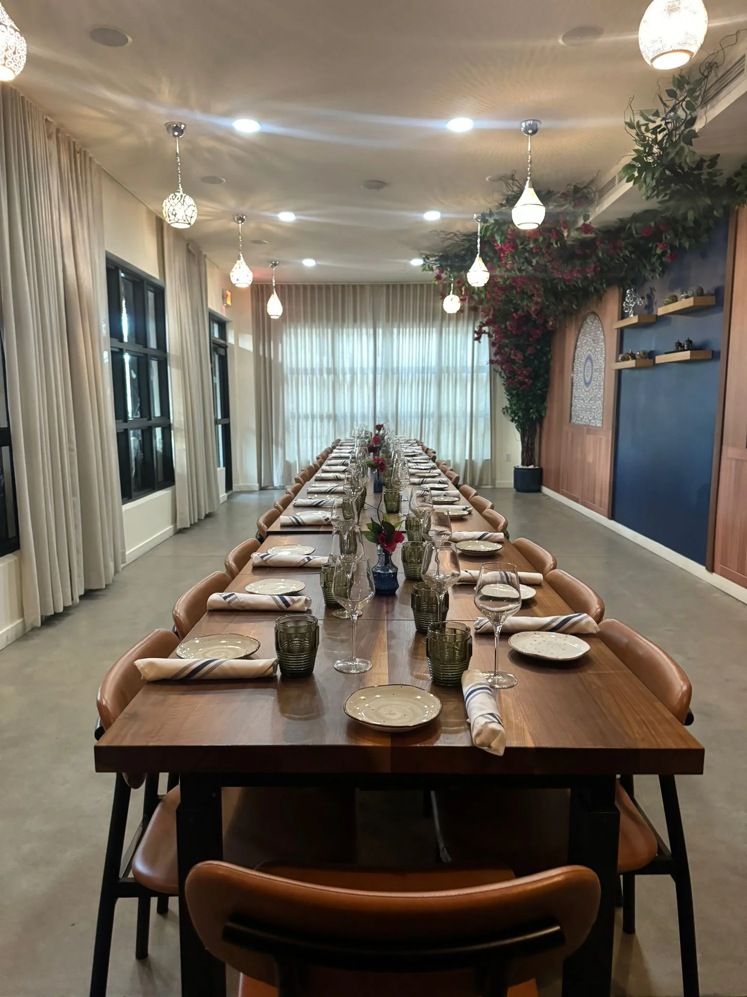 Sufrat private dining room, with dining plate sets, juice and wine glasses with napkins neatly arranged on a long brown wooden table with rows of brown wooden dining chairs on both the right and left side of it.