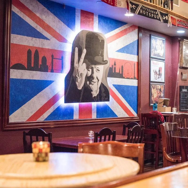 An interior view of a bar featuring wooden tables and chairs, with a mural of a historical figure against a Union Jack backdrop.