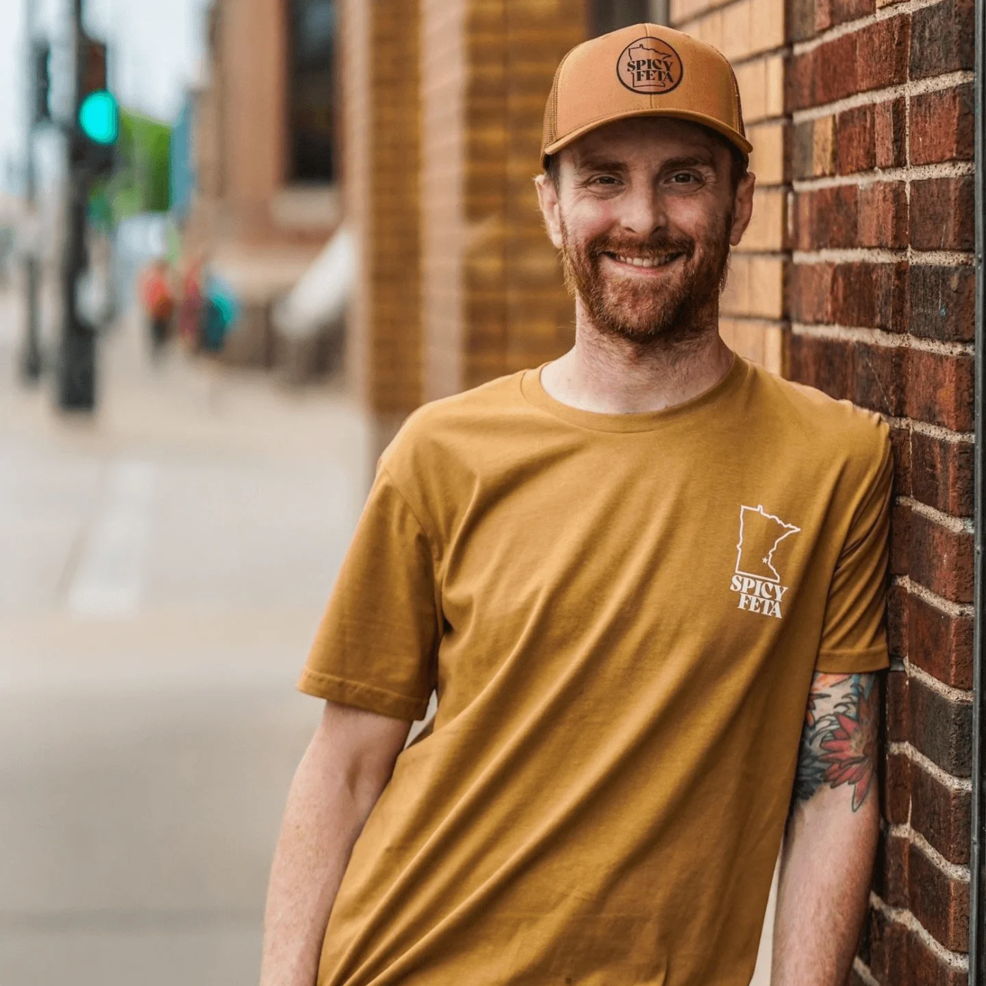 Smiling man in a Spicy Feta t-shirt and cap against a brick wall