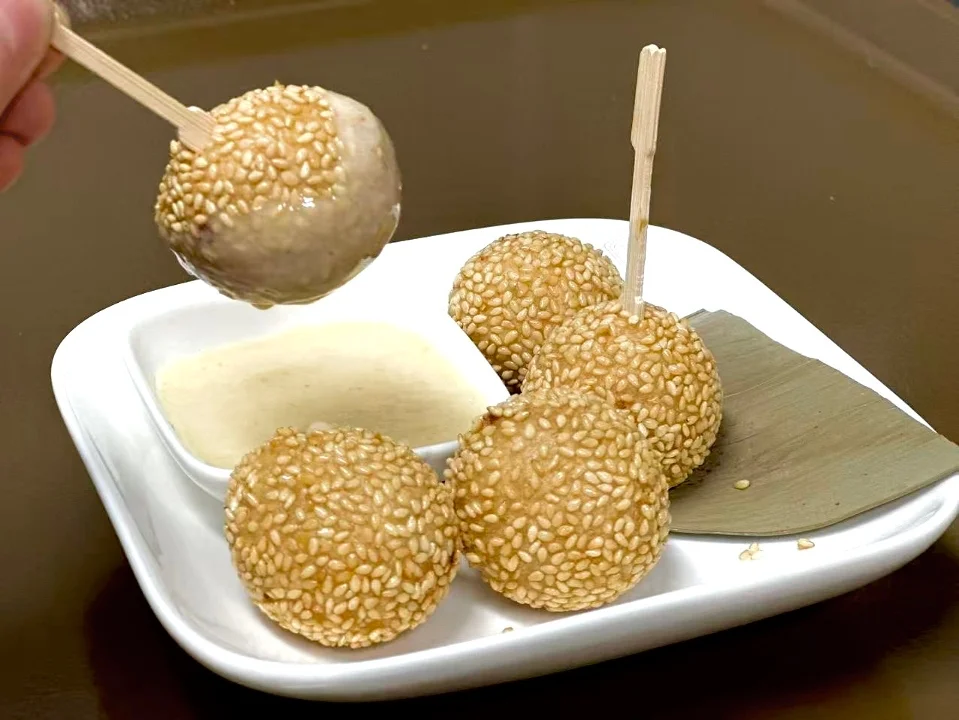 A plate of sesame balls served with a creamy dip. The sesame balls are round and golden-brown, with a hand lifting one on a skewer.