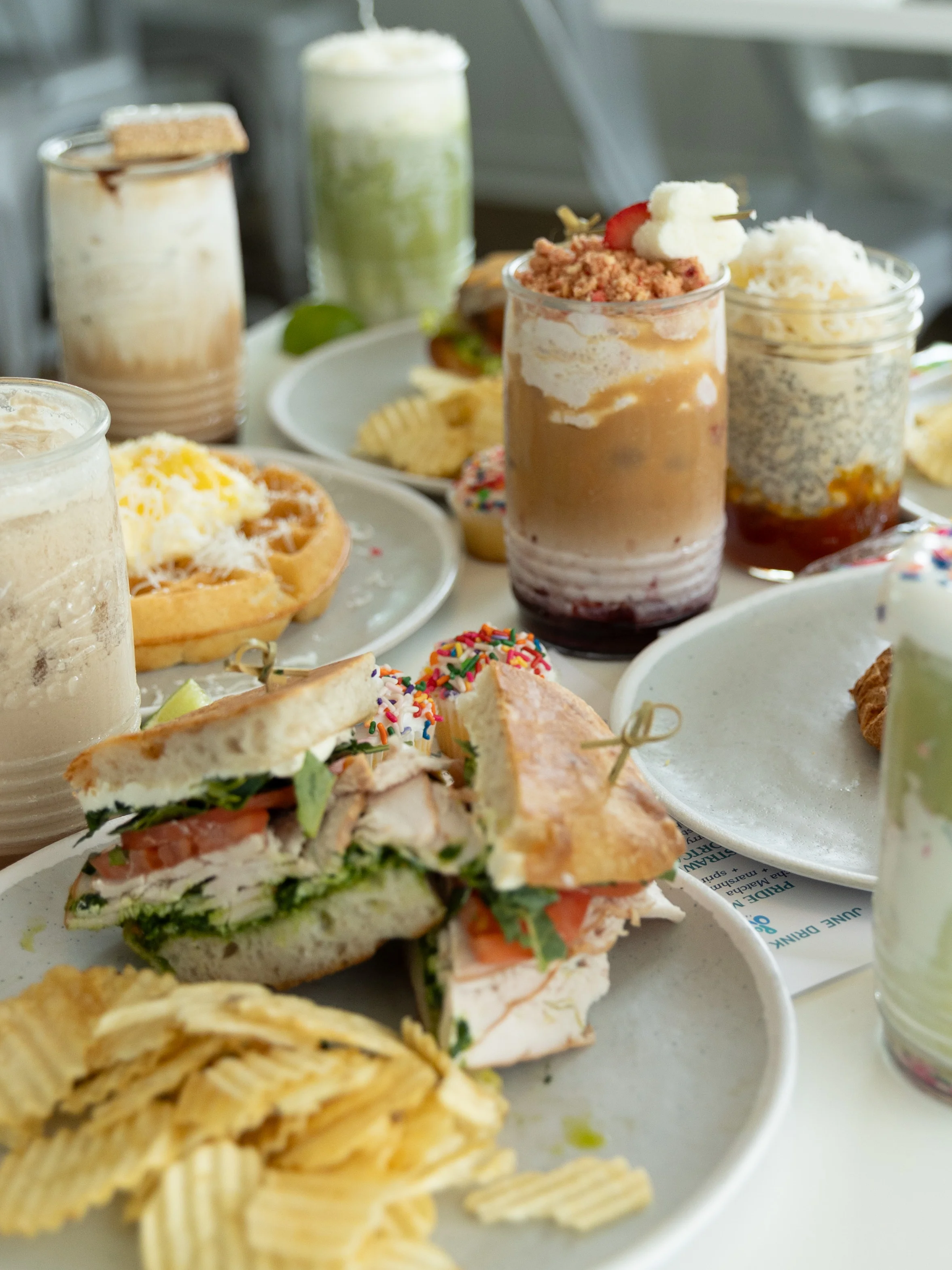 An assortment of food featuring a sandwich filled with greens, tomatoes, and sliced meat, accompanied by several drinks and snacks such as chips and desserts in jars.
